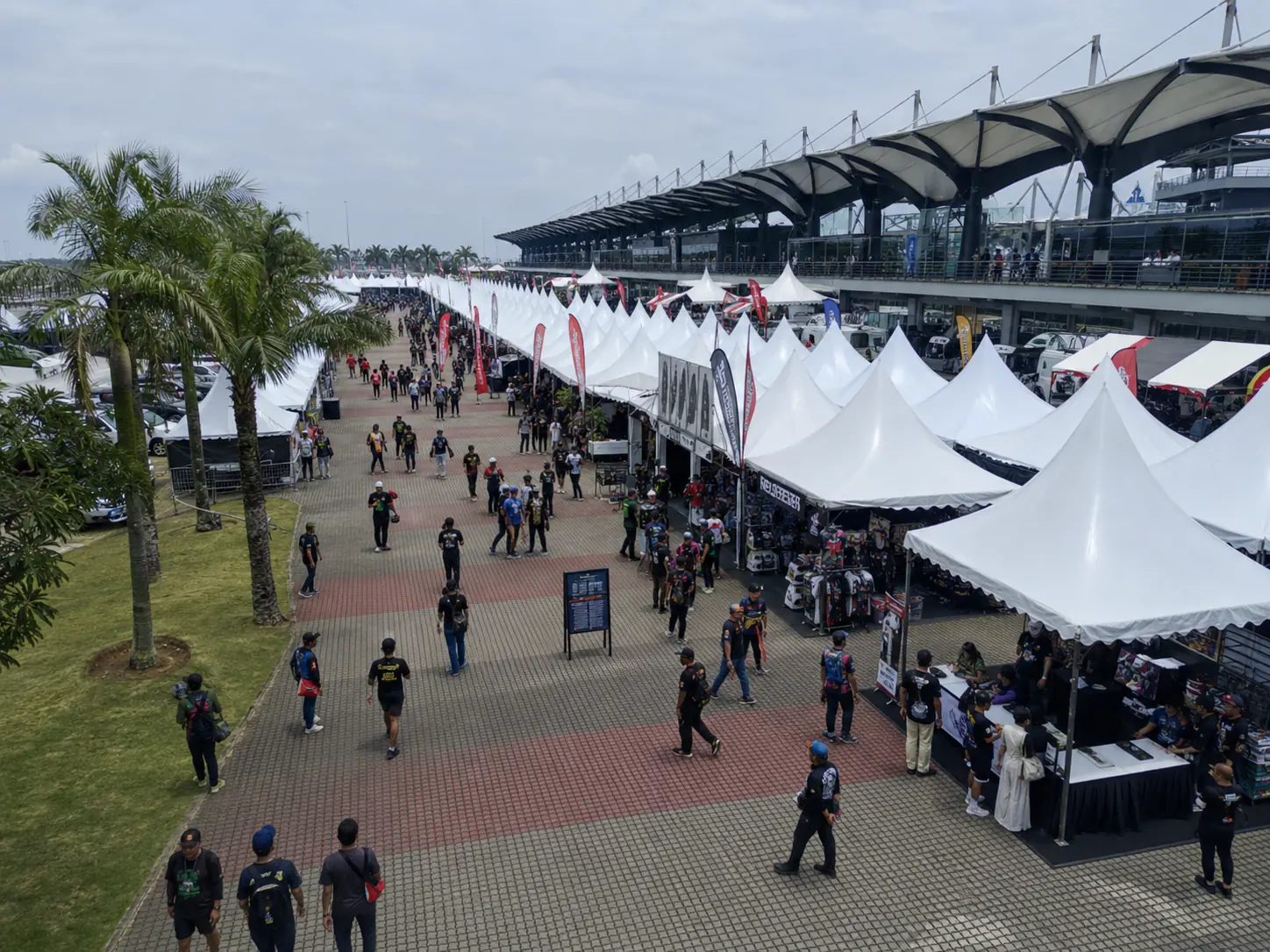 Large outdoor event plaza in Malaysia with long rows of white bazaar canopy tents, vendor booths, event flags and a busy public walkway