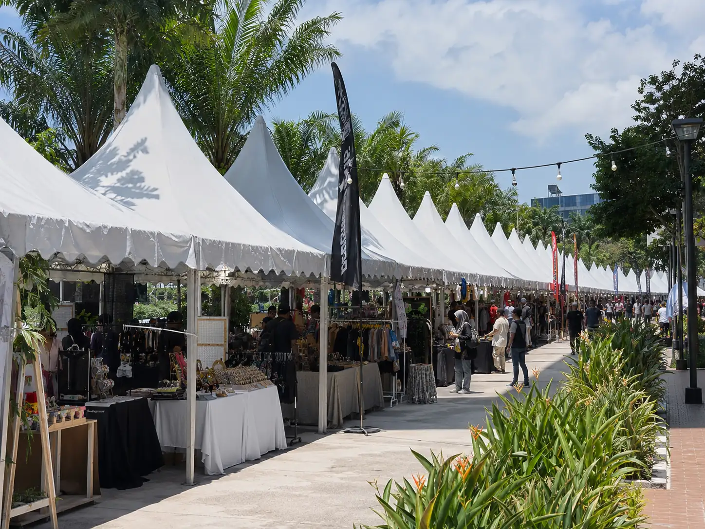 Outdoor bazaar festival scene in Malaysia with white canopy tents lined beside tropical landscaping and market vendors under bright blue skies