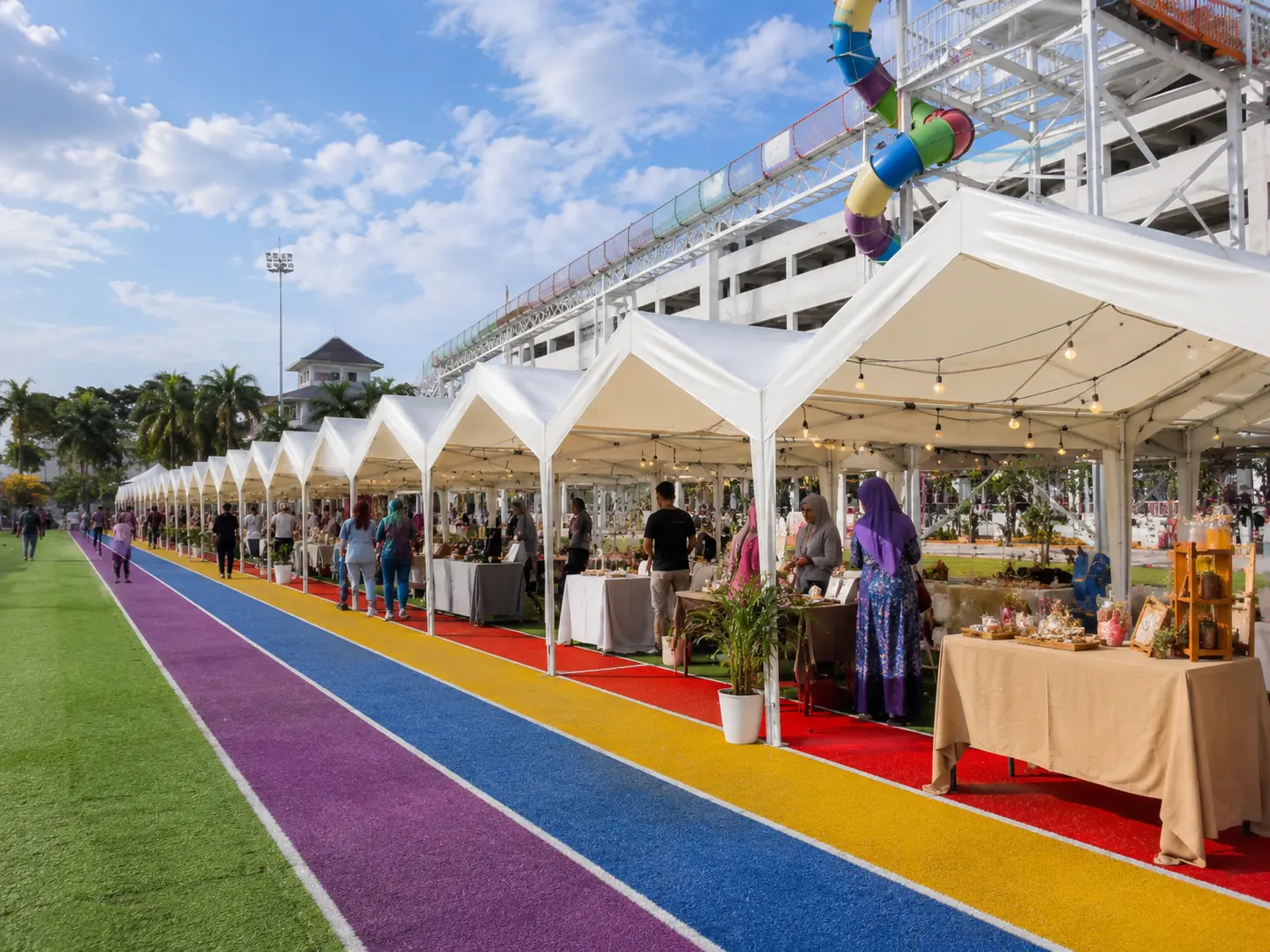 White bazaar tent rental setup in Malaysia beside a colorful sports walkway with lively vendor stalls, shoppers and a bright family event atmosphere
