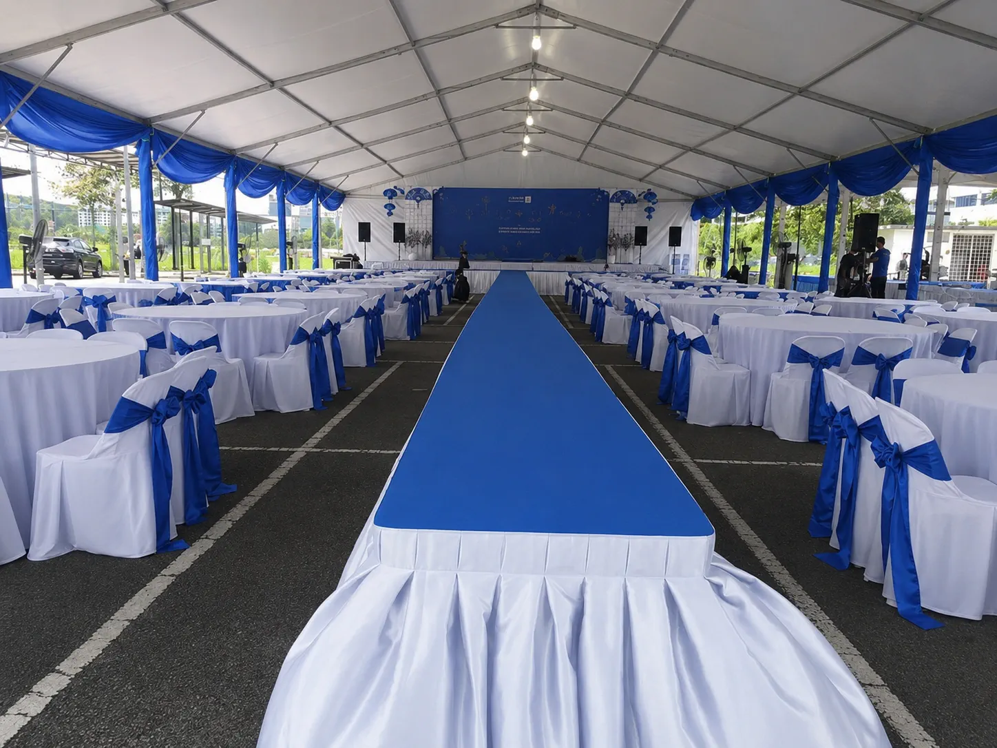 Blue and white banquet marquee setup with central blue stage runway, round dining tables, white chair covers, blue chair ribbons and stage backdrop