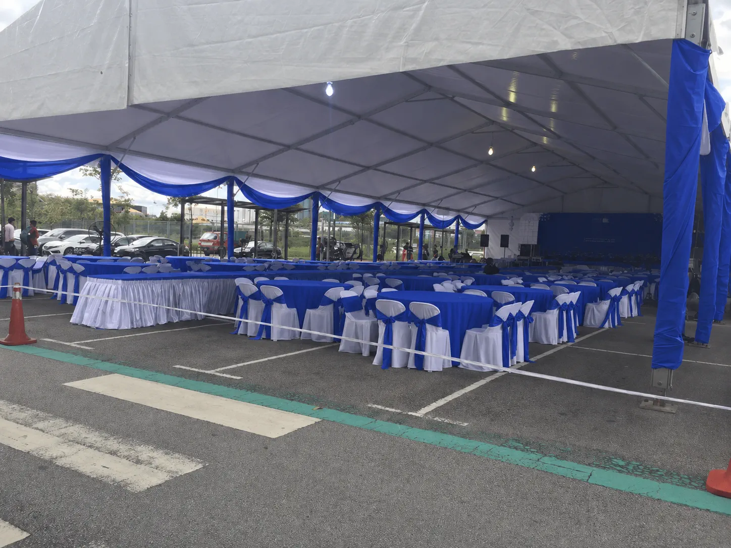 Side view of a blue and white banquet marquee with round tables, blue table skirting, white chair covers, stage backdrop and car park access