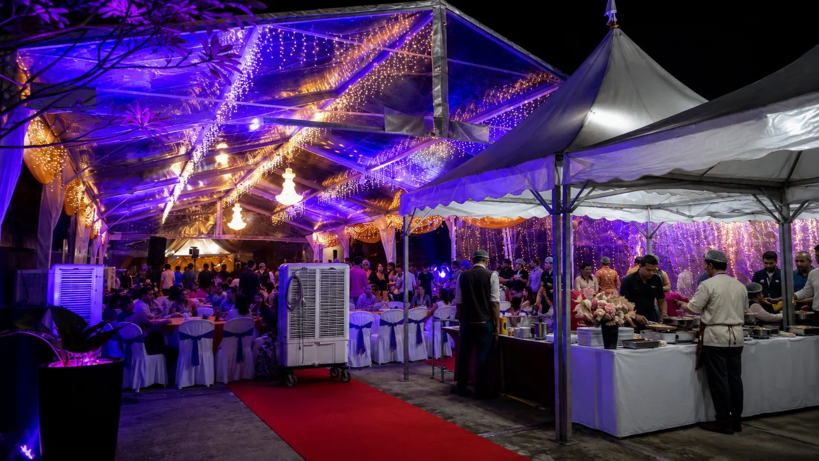 Wedding banquet marquee setup with transparent roof panels, warm fairy lights, dining tables, cooling units, red carpet and covered buffet service tent