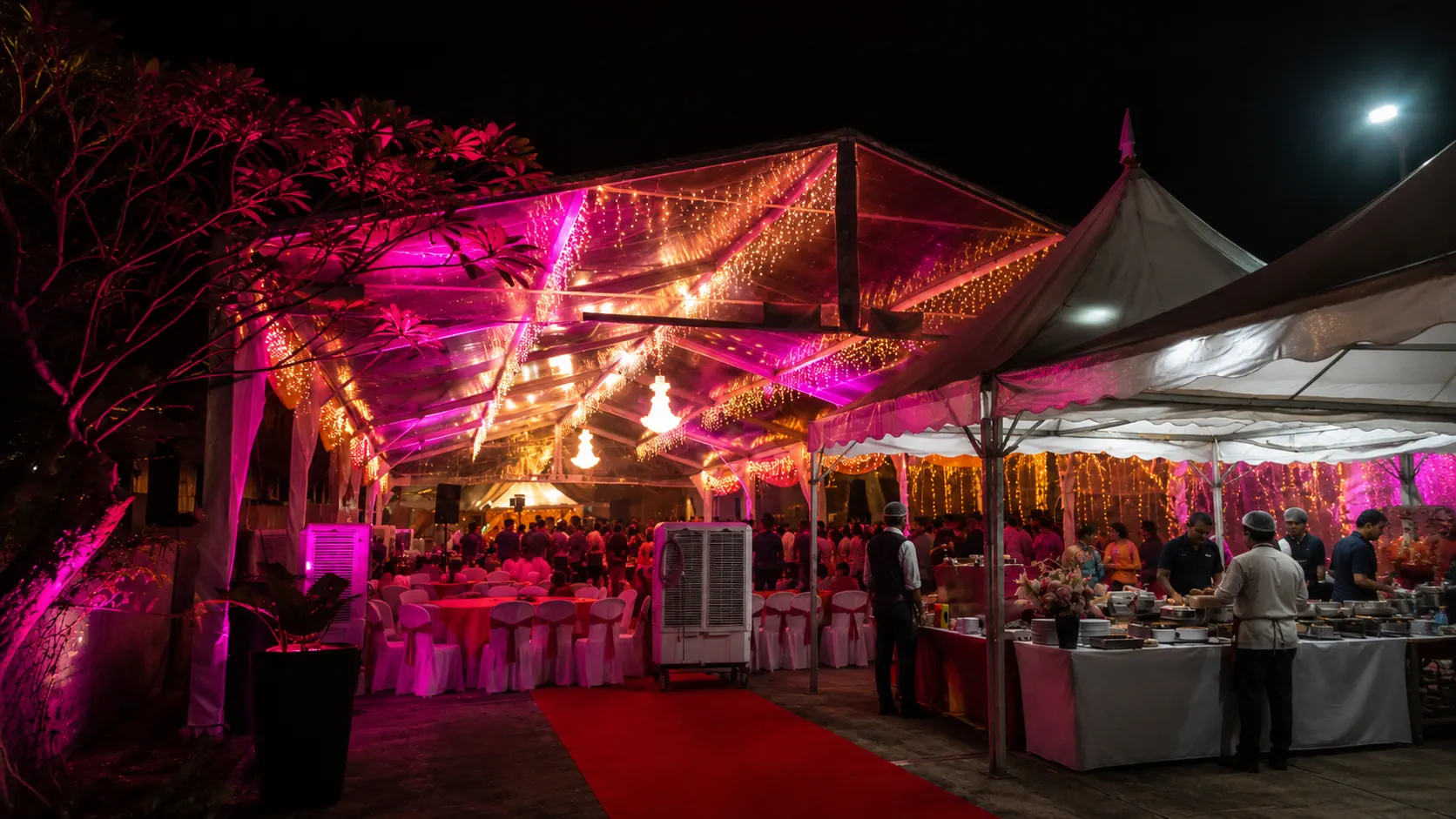 Night wedding banquet entrance with clear roof marquee, pink uplighting, red carpet, white covered chairs, chandelier glow and buffet tent beside the dining area