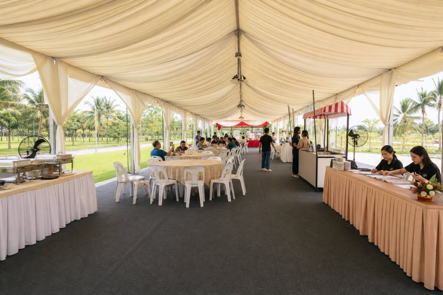 Inside view of an open lawn marquee with dining tables, plastic chairs, registration counter, buffet table, fans and clear walking aisle