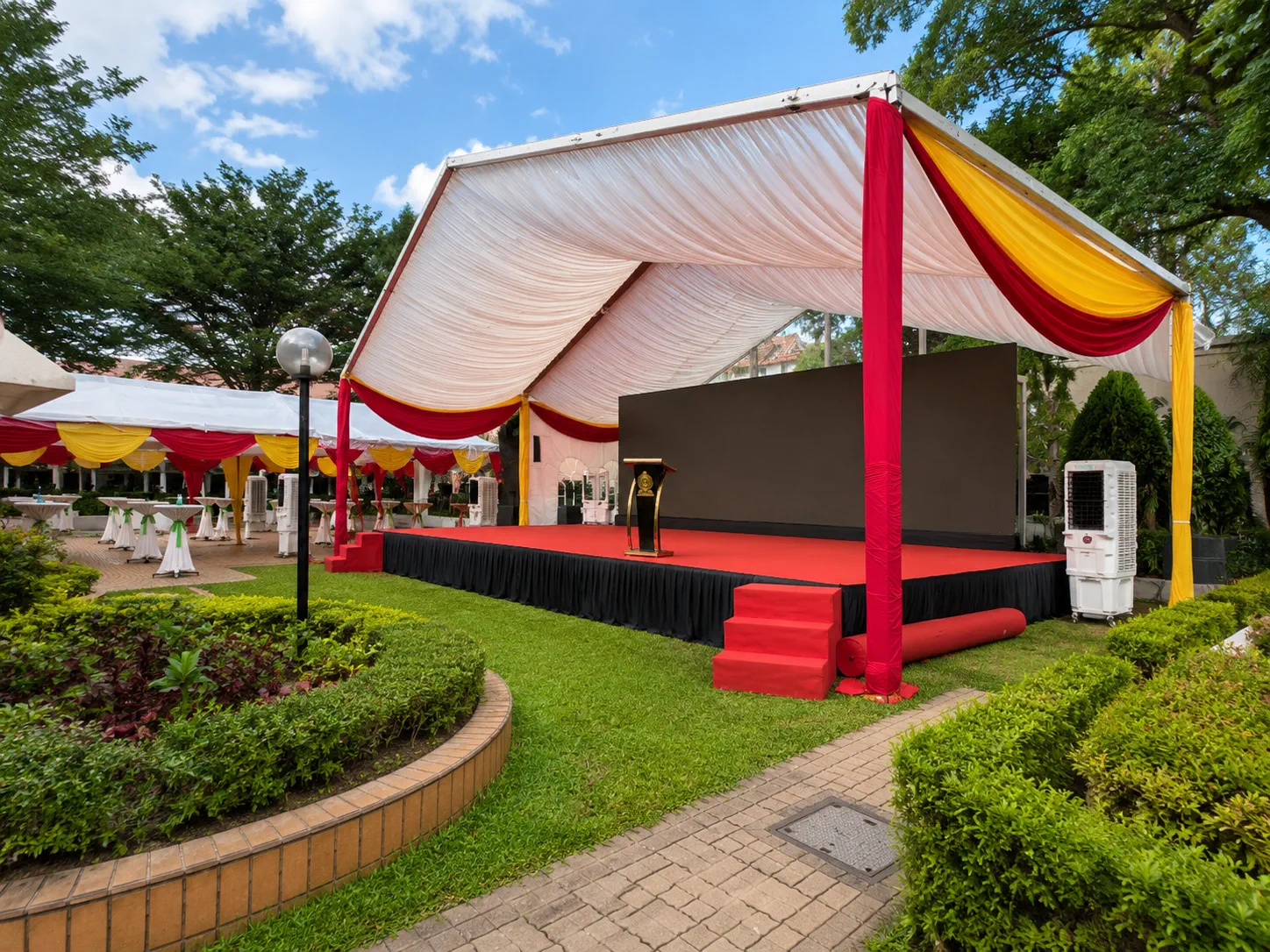 Garden ceremony marquee setup with red and yellow drapes, raised red stage, black backdrop, podium, cooling units and cocktail table area