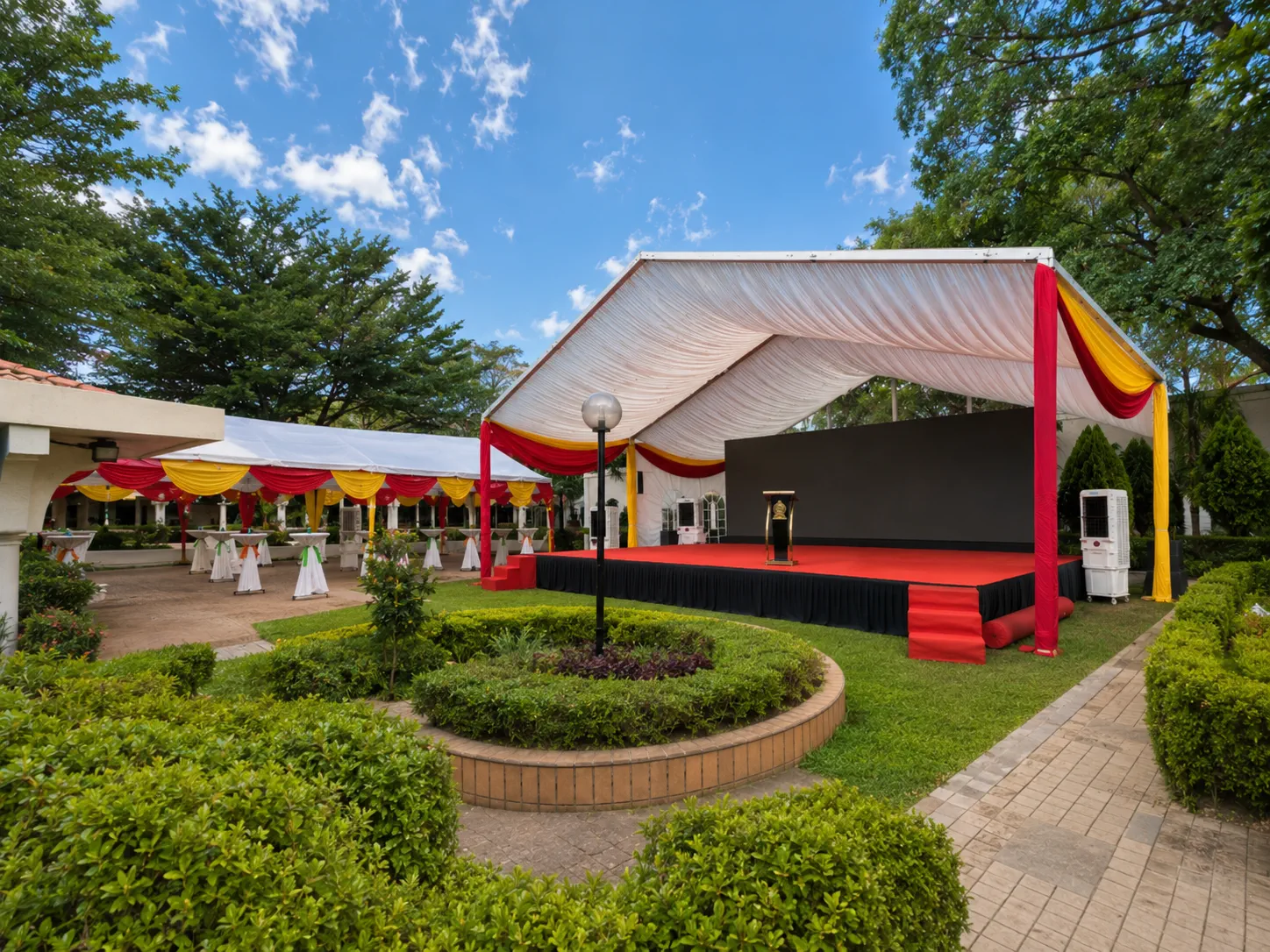 Garden ceremony marquee setup with red and yellow drapes, raised stage, black backdrop, podium, guest canopy, cocktail tables and landscaped courtyard