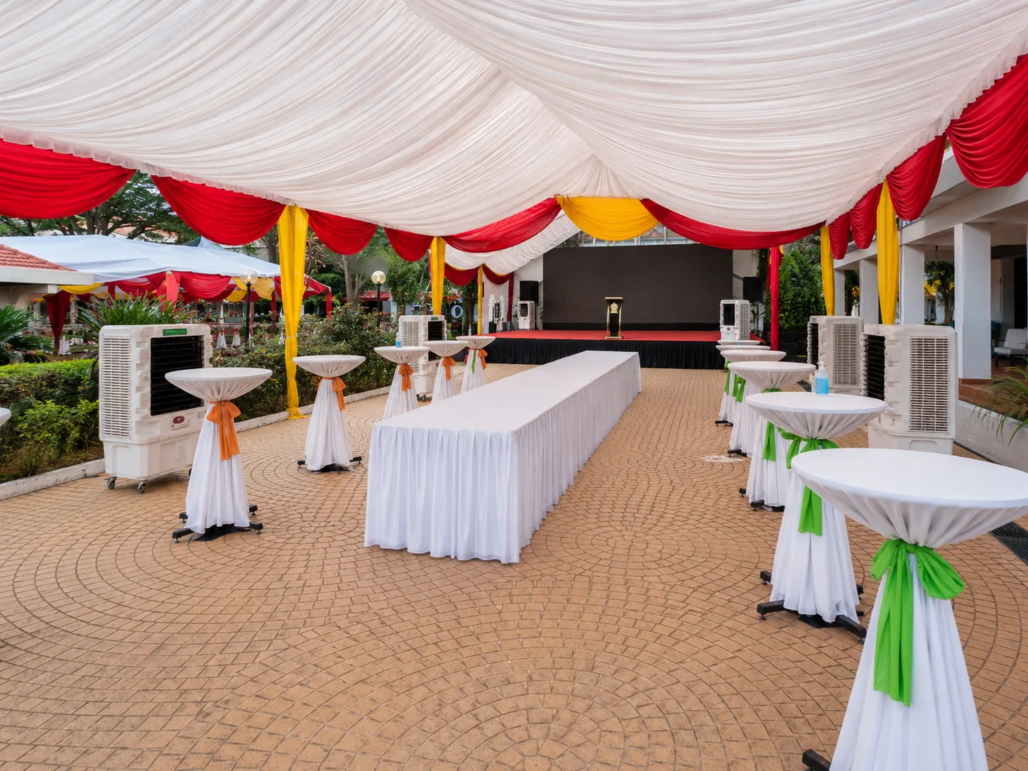 Guest canopy interior with red and yellow drapes, cocktail tables, long buffet table, cooling units and stage view for a garden ceremony
