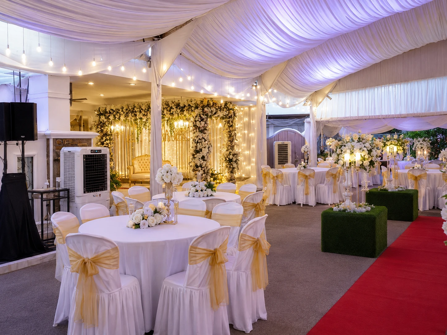 Malay wedding marquee dining setup with bridal stage backdrop, white round tables, gold chair ribbons, red carpet aisle, floral centrepieces and cooling unit