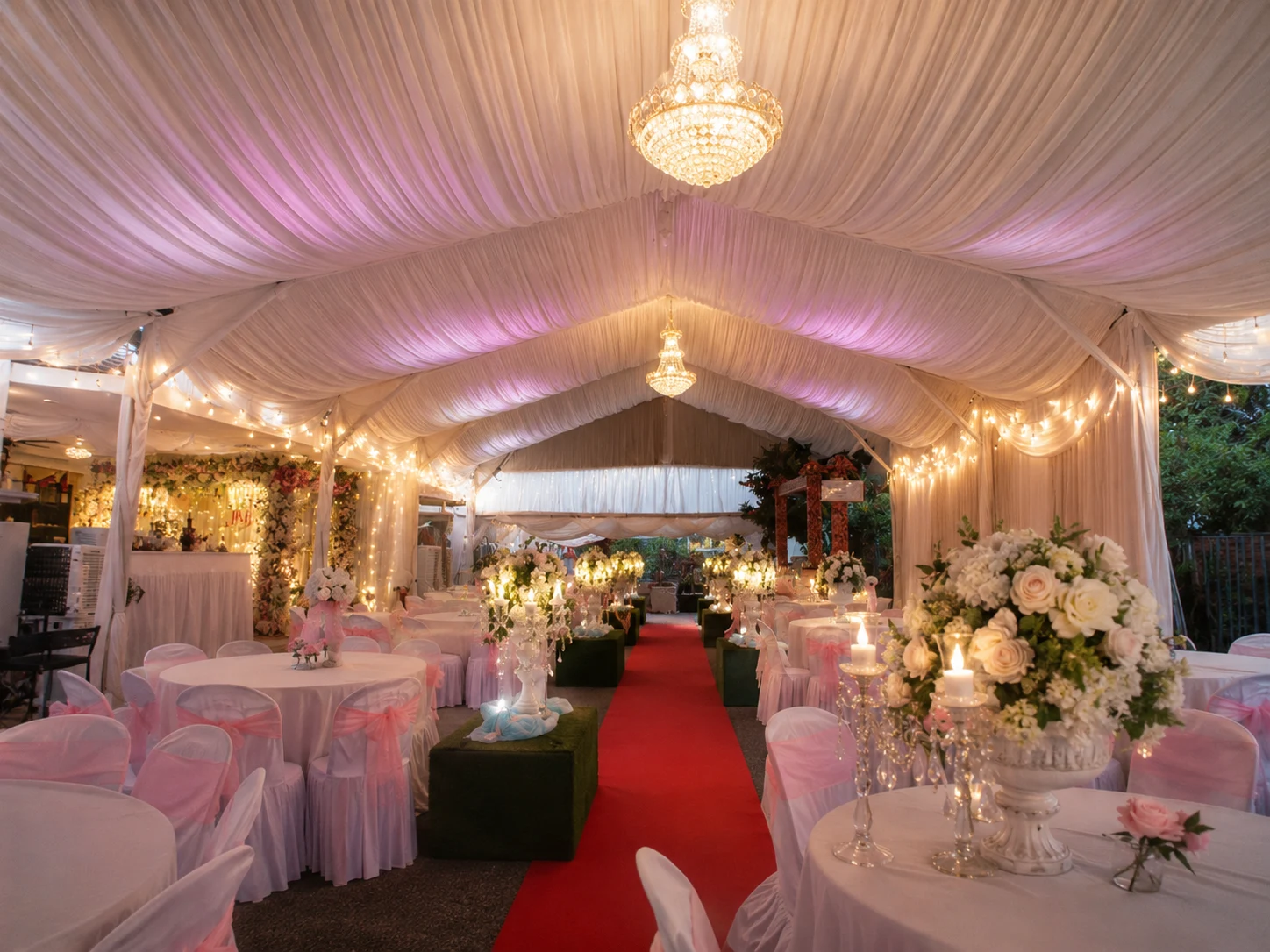 Malay wedding marquee interior with red carpet aisle, chandelier lighting, white ceiling drapes, floral centrepieces and soft pink chair ribbons