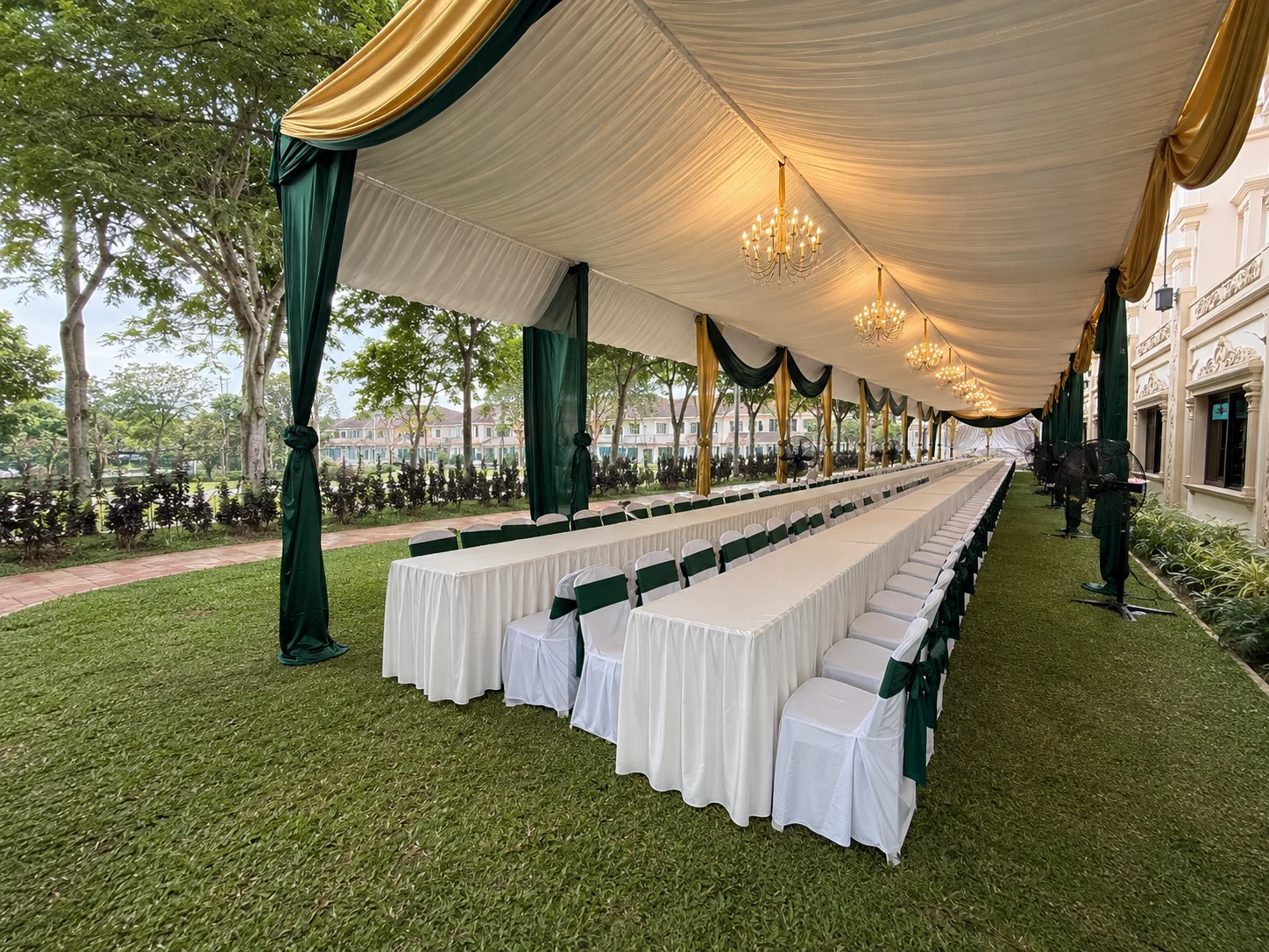 Green and gold long table marquee layout on lawn with chandelier lighting, white chair covers, fans and covered dining rows