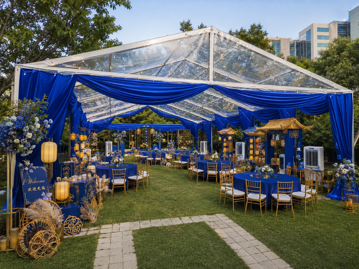 Daytime entrance view of royal blue clear roof marquee with blue fabric swags, gold chairs, round tables, lantern displays, cooling units and garden pathway