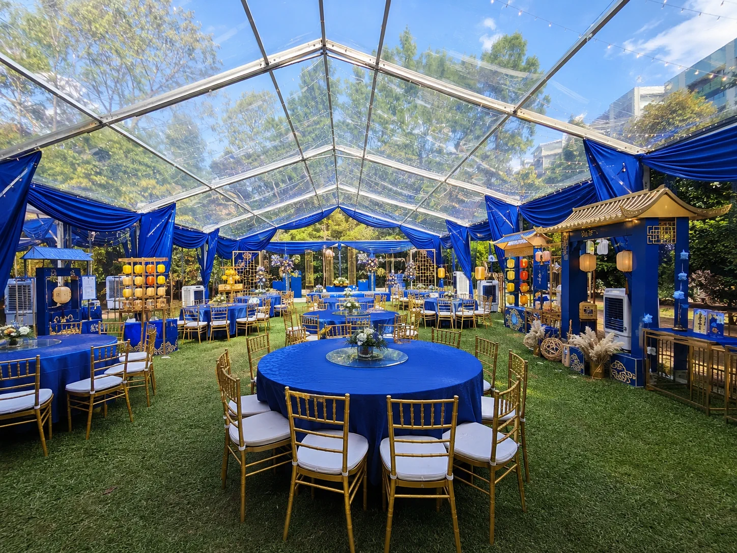 Clear roof banquet marquee with royal blue drapes, gold chairs, round dining tables, transparent roof panels, lantern decor and garden lawn setup