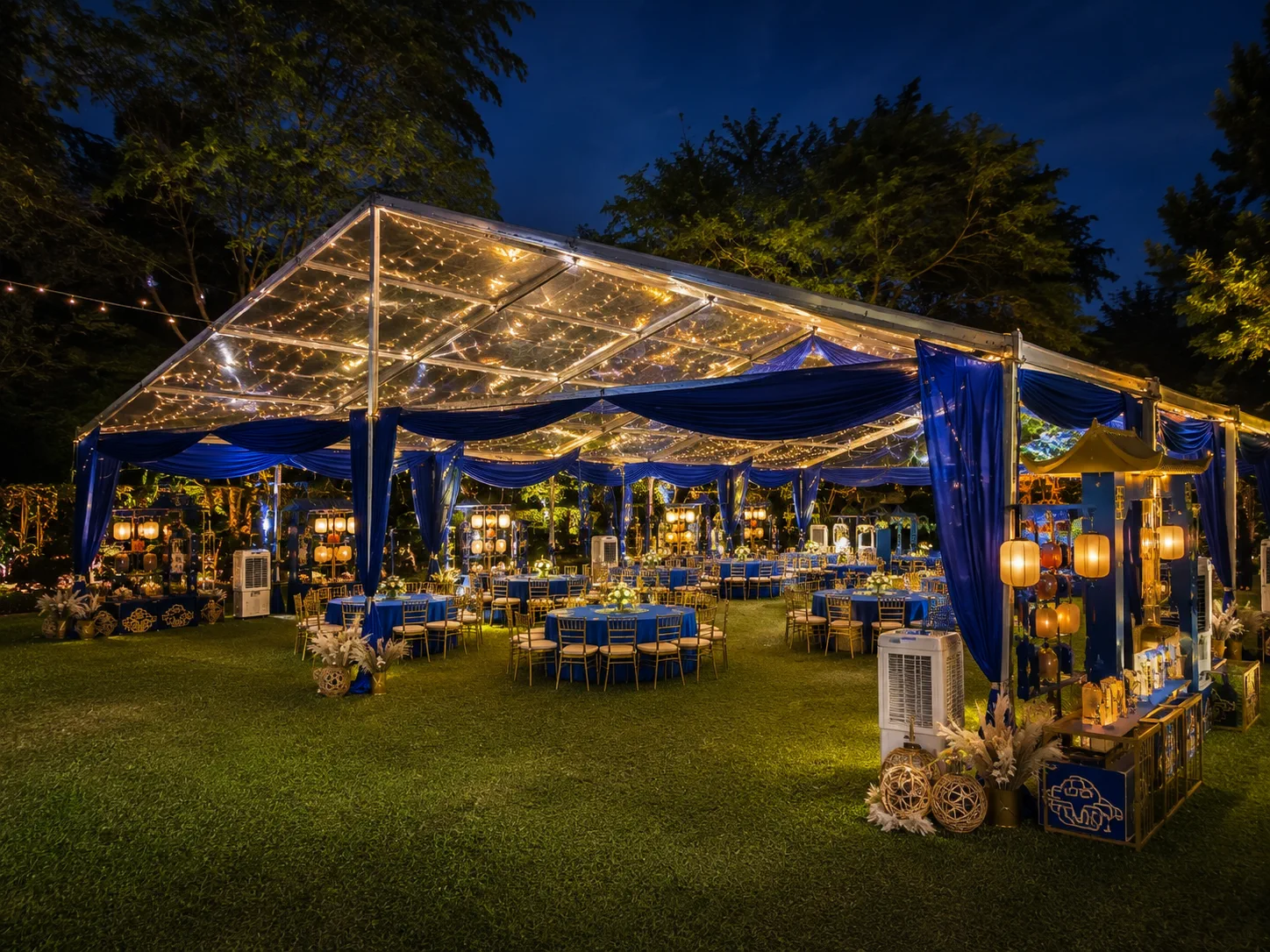 Night view of royal blue clear roof banquet marquee with warm fairy lights, lantern decor, round dining tables, gold chairs and garden lawn lighting