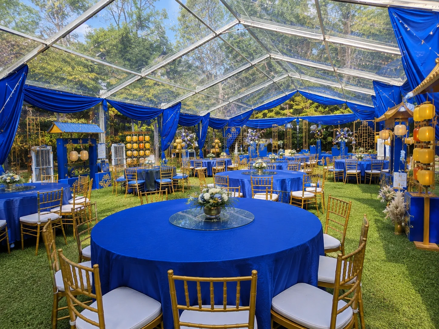 Interior view of royal blue clear roof banquet marquee with round blue tables, gold chairs, floral centrepieces, transparent roof panels and garden backdrop