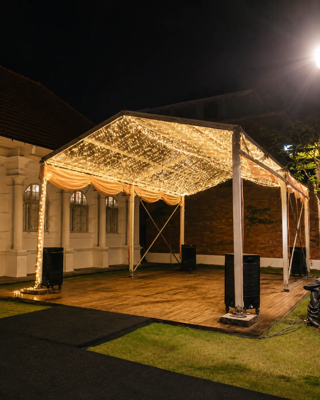 Corner view of a small backyard marquee with clear roof panels, warm fairy lights, timber floor, soft drapes and open house compound layout