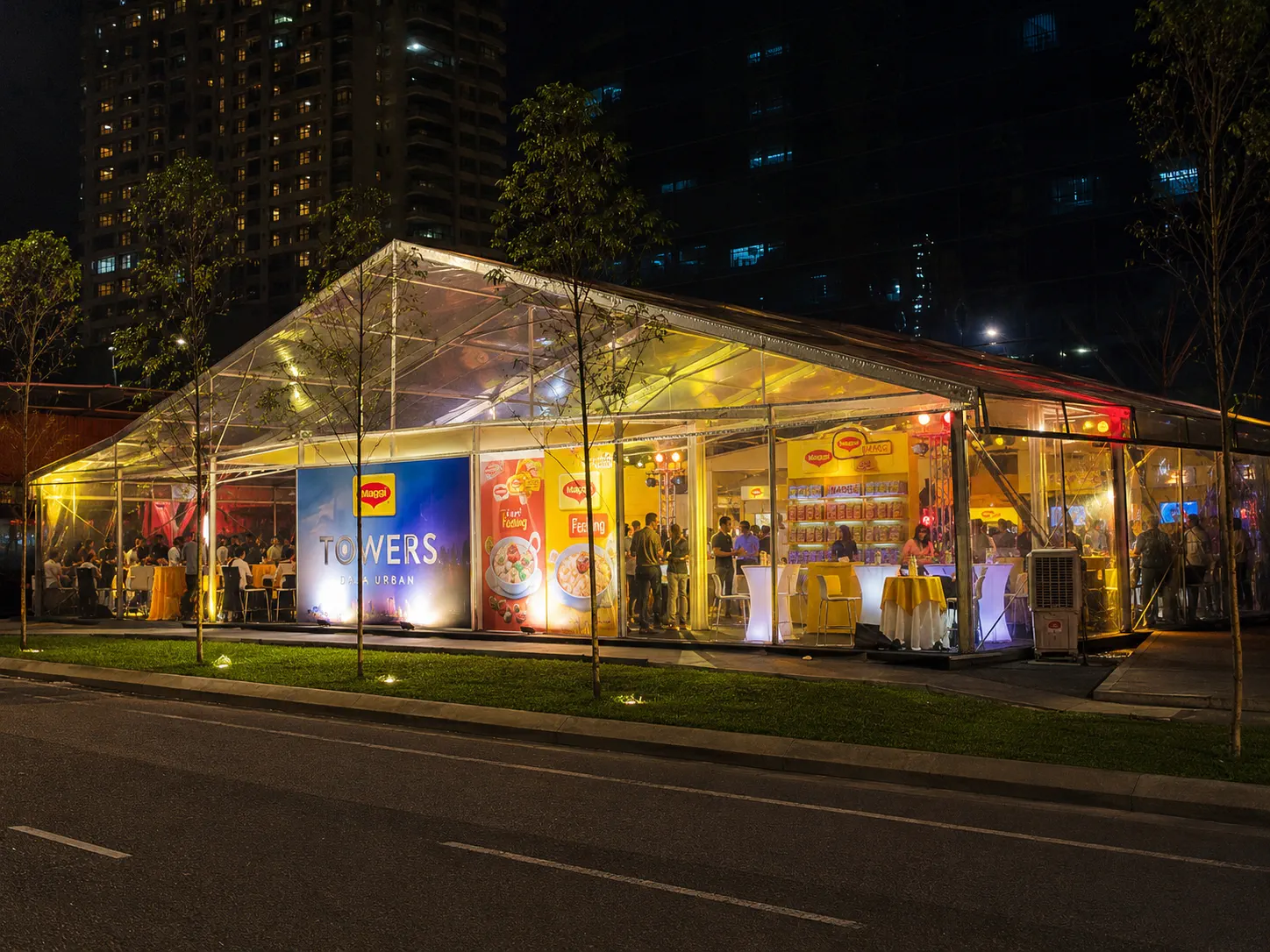 Night exterior view of a clear roof product activation marquee with brand walls, cocktail tables, lighting, clear side panels and city towers