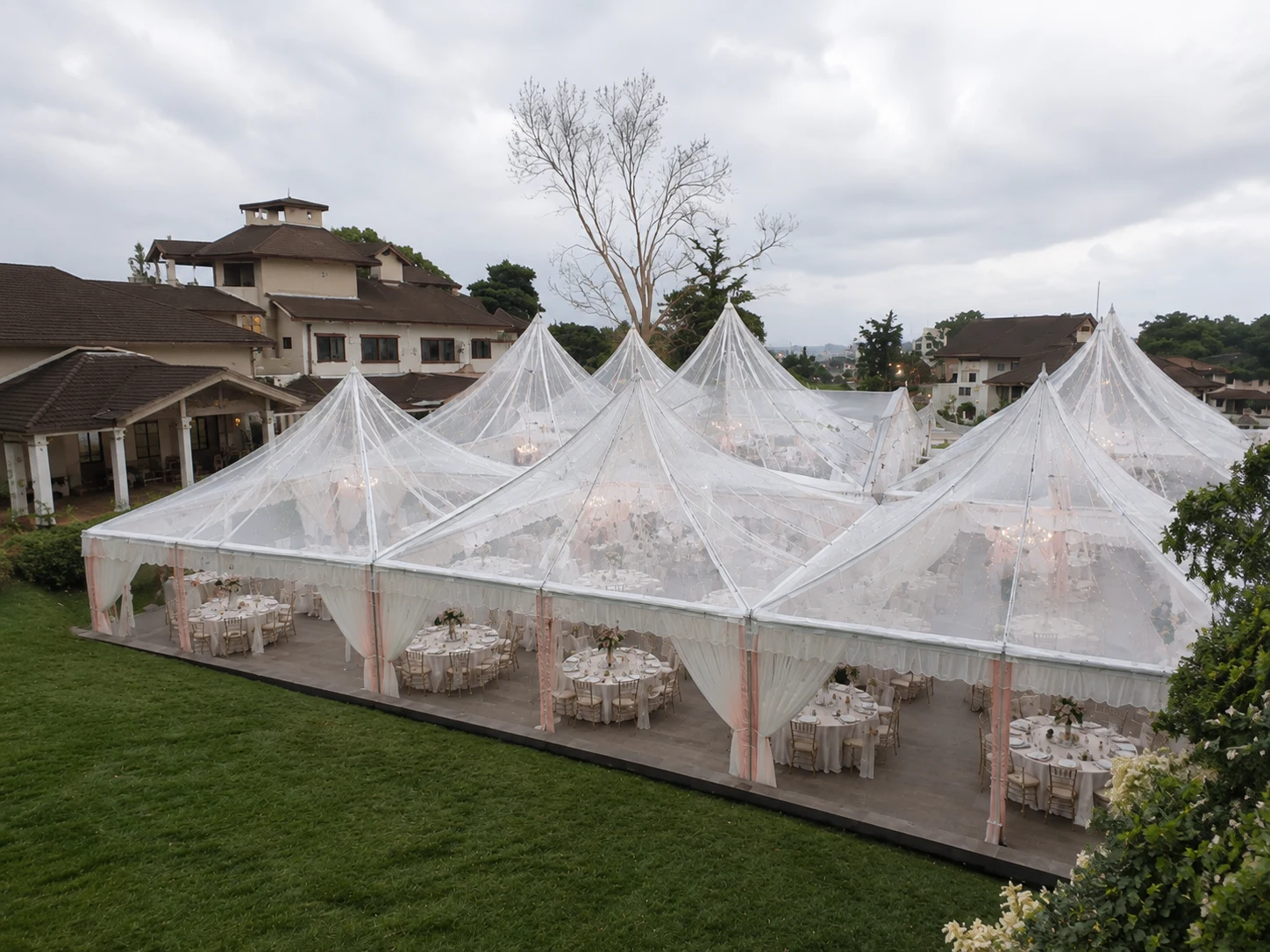 Daytime transparent Arabian canopy wedding setup on a garden lawn with clear pyramid roofs, white drapes and round dining tables