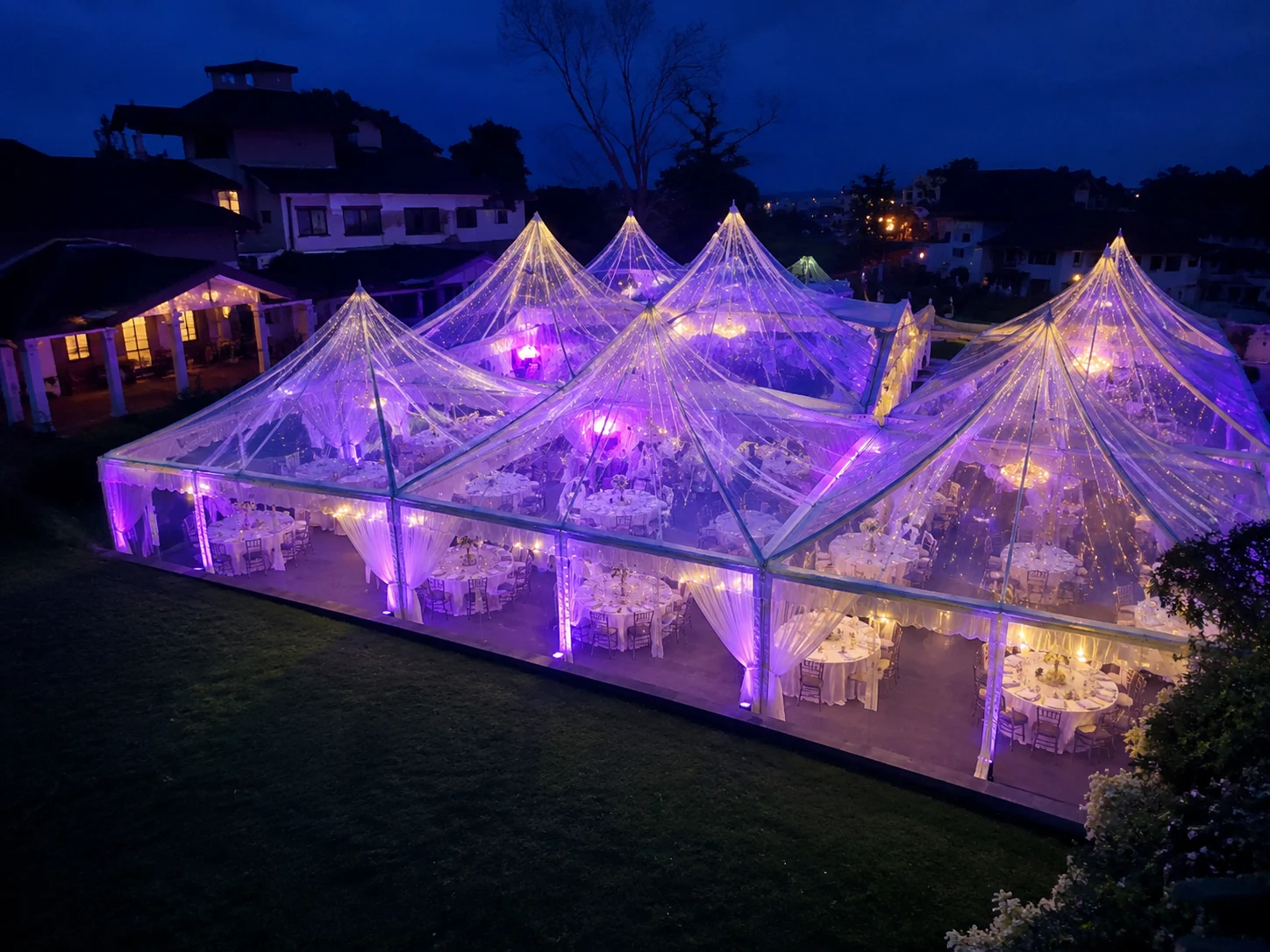 Transparent Arabian canopy wedding setup at night with clear pyramid roofs, purple uplighting, chandelier glow and round banquet tables