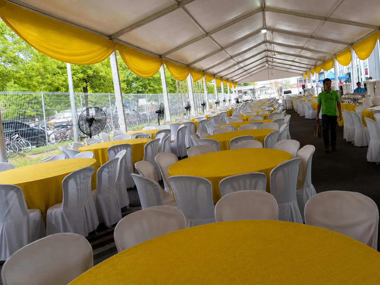 Side view of a large white marquee dining setup with yellow drapes, round banquet tables, white chair covers, standing fans and service aisle